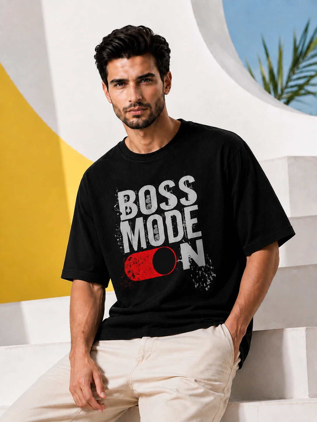 Black oversized T-shirt with "Boss Mode On" typographic print and toggle-switch graphic, front view on male model against a colorful architectural backdrop