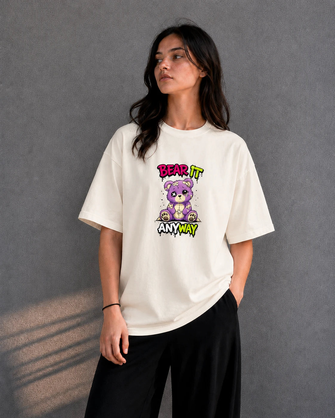Off-white oversized T-shirt with distressed patched teddy bear graphic and "Bear It Anyway" text, front view on female model against grey studio backdrop