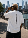 White oversized T-shirt with SHHHH spine typography print, back view on male model on a waterfront promenade with a bridge and skyline