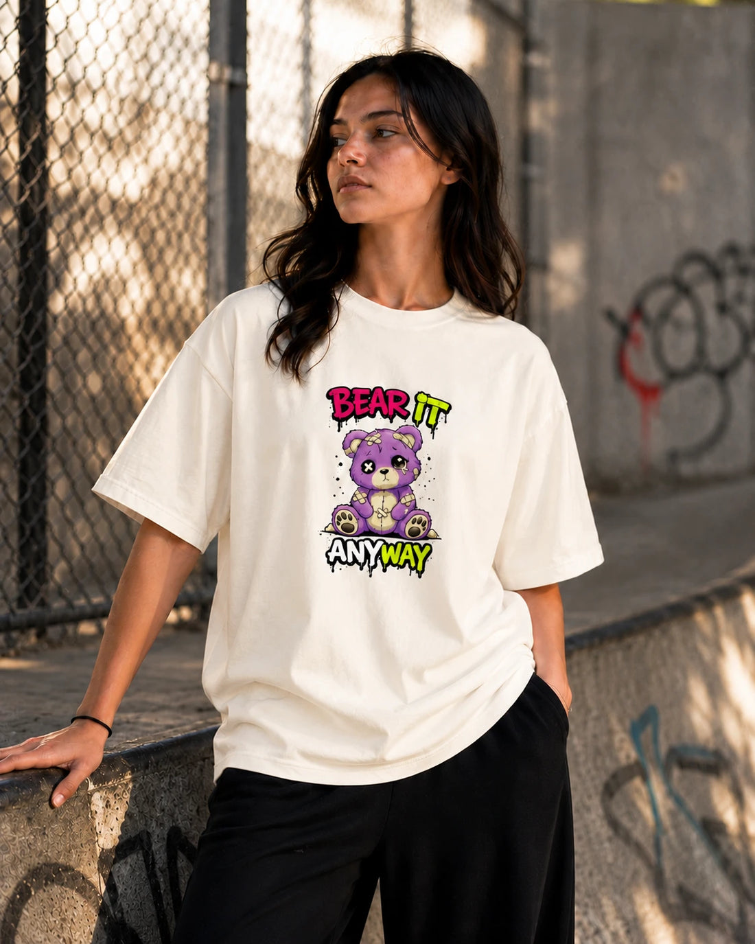 Off-white oversized T-shirt with distressed patched teddy bear graphic and "Bear It Anyway" text, front view on female model against grey studio backdrop