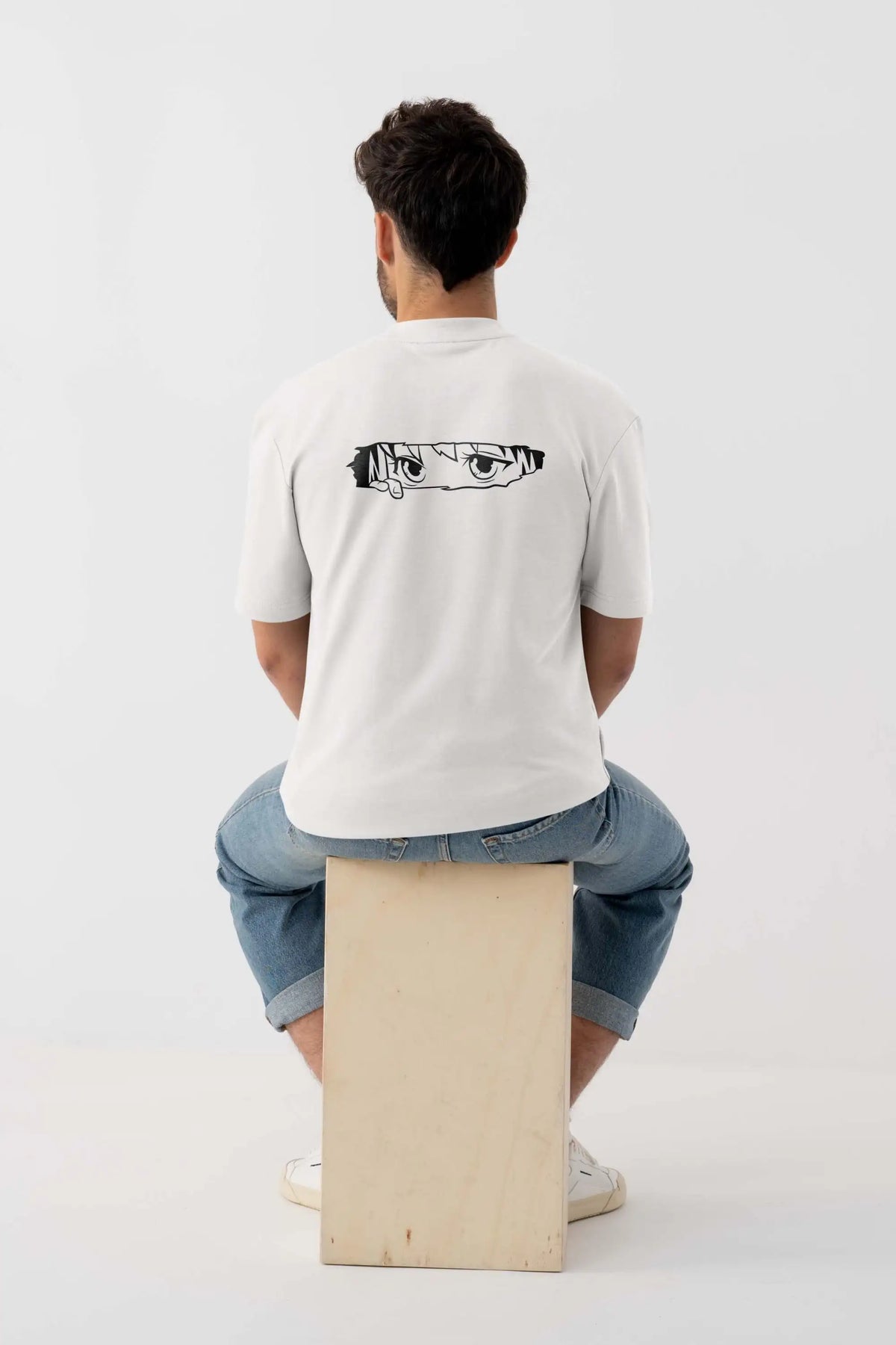 Off-white oversized T-shirt with black-and-white anime eyes graphic print, back view on male model seated on wooden stool